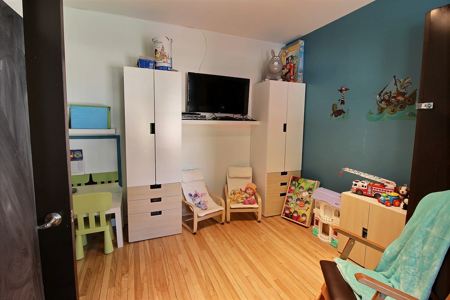 Photo d'une salle de jeux pour les enfants, munie de placards de rangement, d’un bureau et d’une chaise miniatures, de divers jouets, d’une télévision et d’une chaise à bascule.