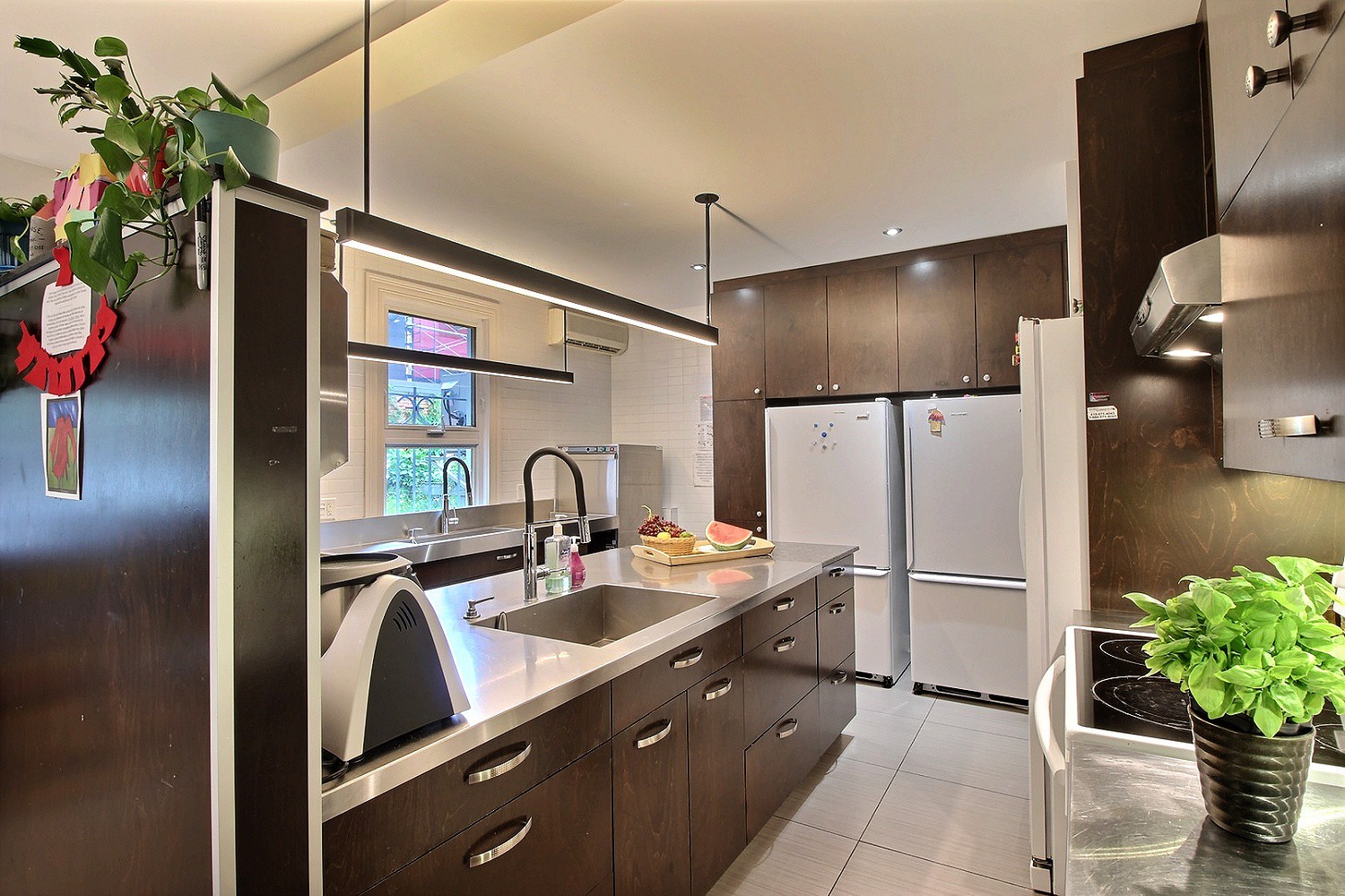 Photo d'une cuisine moderne spacieuse toute équipée (thermomix, plaques de cuisson, four, frigos, plan de travail…) et munie d’une fenêtre.
